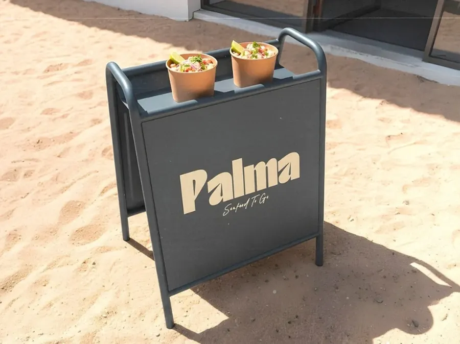 Letrero de acera de Palma Seafood To Go sobre la arena, con dos vasos de comida encima, por CBAS
